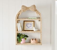 a wicker shelf with books, plants and pictures on it in a white room