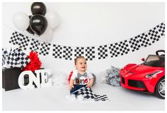 a baby sitting in front of a red sports car and some black and white balloons