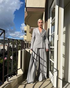 a woman standing on a balcony next to a window wearing a gray suit and matching heels