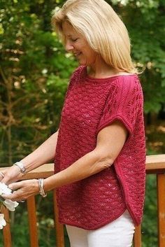 a woman in white pants and a red sweater standing on a wooden deck next to flowers