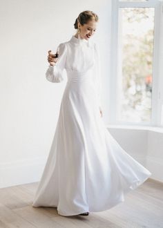 a woman in a long white dress is standing near a window and looking at her cell phone