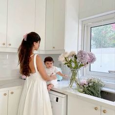 a woman in a white dress standing next to a baby on a kitchen counter with flowers