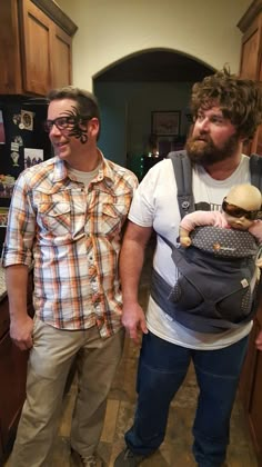 two men standing next to each other in a kitchen holding a baby boy wearing a backpack