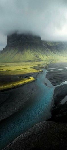 a river running through a lush green field next to a tall mountain covered in clouds