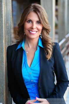 a woman in a blue shirt and black blazer leaning against a wooden fence with her hands on her hips