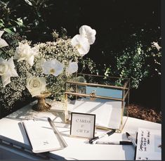 a table topped with white flowers next to a vase filled with water and writing utensils