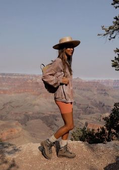 a woman standing at the edge of a cliff with her back to the camera and wearing a hat