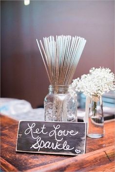 a wooden table topped with two vases filled with white flowers and sticks next to a sign that says let love sparkle