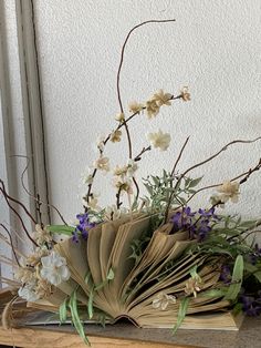 an open book sitting on top of a wooden table next to flowers and branches in front of a window