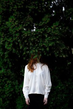 a woman with red hair is standing in front of a green bush wearing a white shirt and black skirt