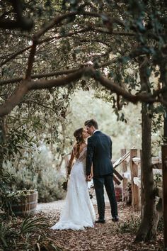 a bride and groom standing under trees in the woods at their outdoor wedding venue, holding hands