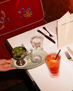 a person holding a plate with olives on it next to a glass and silverware