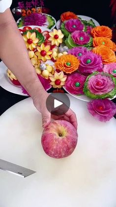 a person cutting an apple on top of a white table with flowers and fruit in the background