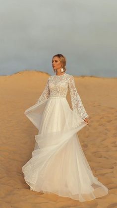 a woman standing in the sand wearing a wedding dress with long sleeves and bell sleeves