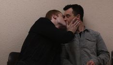 two men are kissing each other while sitting on a chair in front of a wall