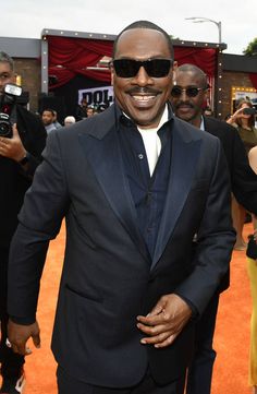 a man in a tuxedo and sunglasses walking on an orange carpet with people behind him