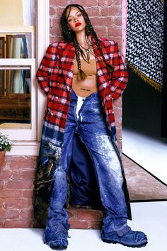 a woman with dreadlocks leaning against a brick wall wearing jeans and a plaid shirt