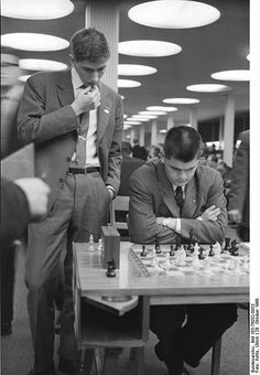 two men are playing chess in an office setting, one is wearing a suit and the other is sitting at a table