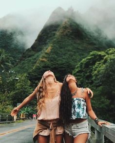 two young women standing on a bridge looking up at the mountains and clouds behind them