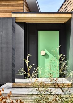 a green door sits in front of a black and wood building with plants growing outside