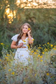 a girl in a white dress standing in a field with yellow flowers and holding a flower