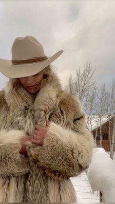 a woman wearing a fur coat and hat standing in the snow with her arms crossed