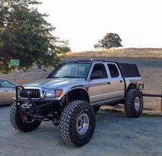 a silver truck parked next to two cars on top of a dirt field with trees in the background