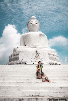 a woman sitting on the steps in front of a large buddha statue with clouds behind her