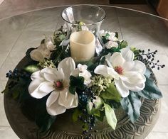 a glass table topped with white flowers and greenery next to a candle on top of it