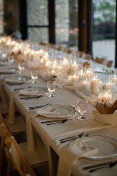 a long table is set with white linens and place settings, lit by candles