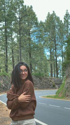 a woman standing on the side of a road with her arms crossed and trees in the background