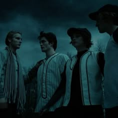 four young men are standing in front of a dark sky and trees, one is wearing a baseball cap