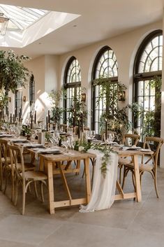 a table set up for a wedding with flowers and greenery on the tables in front of large windows