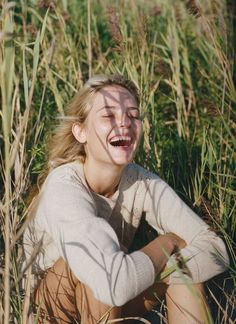 a woman sitting in tall grass with her eyes closed and mouth wide open, laughing