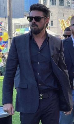 a man wearing sunglasses and a suit walking in the grass with other people behind him