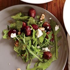 a white plate topped with salad next to a bowl filled with fruit and cheese on top of a wooden table