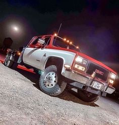 a red and white truck parked on top of a dirt field at night with its lights on