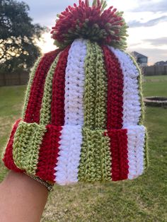 a hand is holding up a crocheted hat in the grass with red, white and green stripes