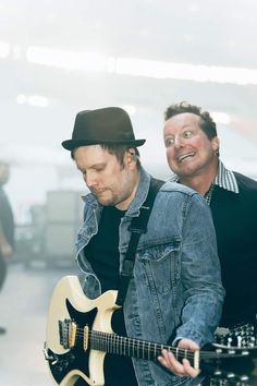 two men are playing guitars in an indoor area, one is wearing a denim jacket and the other has a black hat