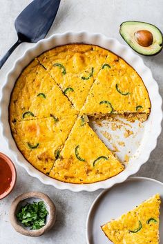 a quiche with one slice missing from it on a plate next to an avocado