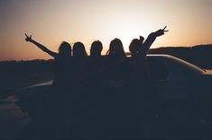 three girls standing in front of a car with their arms up at the sun set