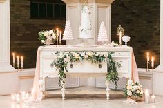 a table with candles and flowers on it in front of a white wedding cake decorated with greenery