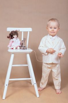 a little boy in a suit standing next to a chair with a doll on it