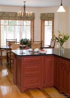 a kitchen with wooden floors and red cabinetry, lights on the windows above the island