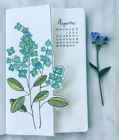 an open notebook with blue flowers on it next to a flower marker and some green leaves