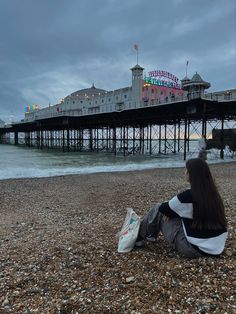 a woman sitting on the beach looking out to sea with a pier in the background