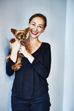 a woman is holding a small dog in her arms and smiling at the camera while standing against a wall