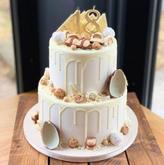 a three tiered cake with white frosting and gold decorations on top is sitting on a wooden table