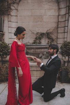 a man kneeling down next to a woman in a red dress and black suit on the ground