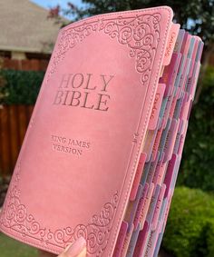 a hand holding a pink bible in front of a house with trees and bushes behind it